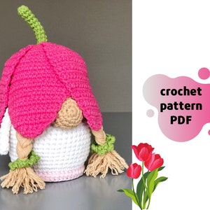 May include: Crochet pattern PDF for a pink and white gnome with a flower hat and braided hair. The gnome is sitting on a white surface. The text "crochet pattern PDF" is in a pink and white blob shape.