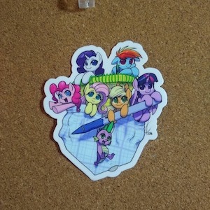 May include: A sticker featuring a group of cartoon ponies and a dragon, all in a diaper. The ponies are pink, purple, blue, yellow, and orange. The dragon is green. The diaper is blue and white with a grid pattern. The sticker is on a brown corkboard.
