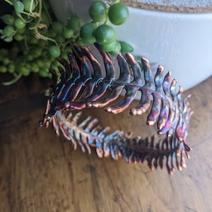 May include: A copper fern leaf bracelet with a hammered finish and iridescent blue and purple hues.