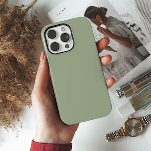Peut inclure: Une coque de smartphone vert sauge uni avec une finition mate, dotée d'une découpe rectangulaire pour l'appareil photo avec trois objectifs circulaires et un flash, conçue pour un appareil mobile moderne aux coins arrondis.
