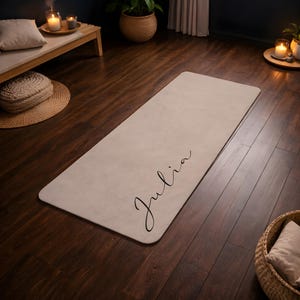 May include: A rectangular beige yoga mat featuring the name Julia written in a black cursive script font. The mat has rounded corners and a smooth, solid-colored surface, positioned flat on a dark wooden floor in a room with soft lighting.