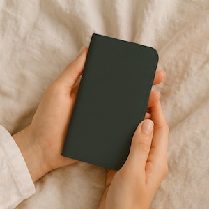 Puede incluir: Una funda de teléfono de cuero verde oscuro sostenida en dos manos. La funda tiene una superficie texturizada y un borde cosido. El fondo es una tela beige claro.