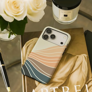 May include: A smartphone case with a wave design in shades of beige, peach, brown, and teal. The case is lying on a book with the word "ART" visible. A black and gold pen, a candle, and white roses are also in the image.