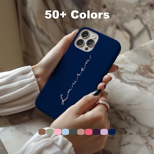 Puede incluir: Una funda de teléfono azul marino con el nombre "Lauren" en escritura blanca. La imagen muestra una variedad de opciones de color debajo de la funda, con el texto "50+ Colors" en la parte superior. La funda está en manos de una persona.