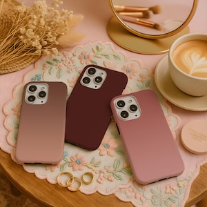 May include: Three phone cases in shades of pink and burgundy are arranged on a floral embroidered cloth. A gold-framed mirror, makeup brushes, and a cup of coffee with latte art are in the background. Gold rings and a small makeup compact are also visible.