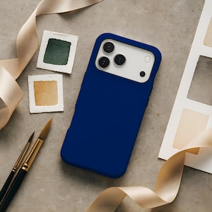 May include: A navy blue phone case with a white camera panel. The case is surrounded by art supplies, including paint swatches and paintbrushes. A cream-coloured ribbon is draped around the items.