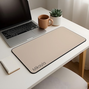 May include: A rectangular beige desk mat with a black border and the name Mikayla printed in black in the bottom left corner. The mat is positioned on a white surface next to a silver laptop, a brown ceramic mug, a small succulent, and a notebook.