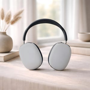 May include: White and black over-ear headphones with a black headband and silver accents. The earcups are white with black trim. The headphones are resting on a light-colored surface, with a vase and books in the background.