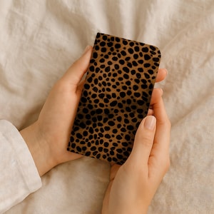 Puede incluir: Una funda rectangular con un estampado de leopardo marrón. La funda se sostiene con dos manos sobre un fondo neutro. La funda tiene un patrón repetido de manchas marrones oscuras.