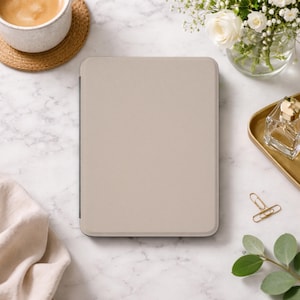 May include: A beige tablet case with a textured surface. The case is rectangular and has a dark gray border. The image also includes a cup of coffee, flowers, and a gold tray with a perfume bottle.