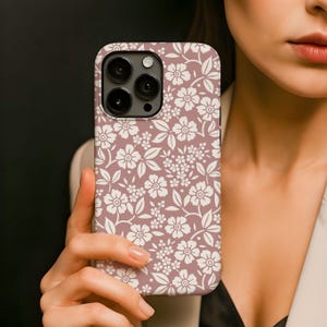 Pode incluir: Uma capa de telemóvel com um padrão floral. A capa tem um fundo malva com um padrão repetido de flores e folhas brancas. A capa do telemóvel tem um recorte para a câmara com três lentes.
