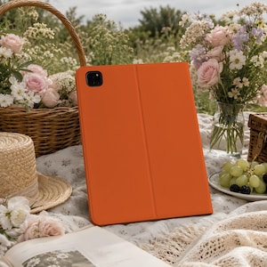 May include: An orange tablet case with a textured finish is open, revealing the screen. The case is on a picnic blanket with a straw hat, flowers, and a basket in the background. The scene is outdoors.