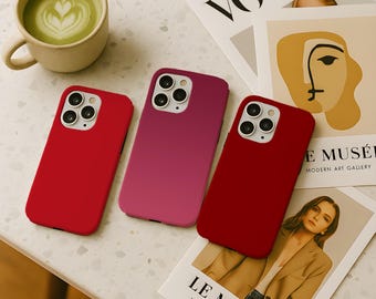 May include: Three phone cases in red and pink hues are displayed on a speckled surface, alongside a cup of matcha latte and fashion magazines. The cases have multiple camera lenses. The magazines have the words "VOGUE" and "LE MUSÉE" printed on them.