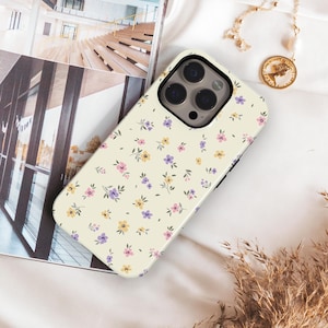 May include: A cream-coloured smartphone case featuring a repeating pattern of small, delicate flowers in shades of pink, yellow, and purple with green leaves. The case has a large cutout for a triple-lens camera system on the back.