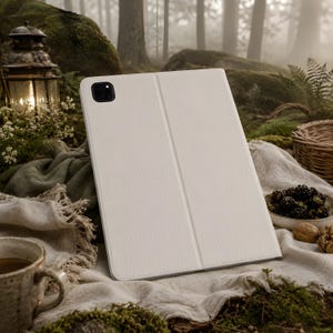 Op de afbeelding: Een witte tablet hoes met een getextureerd oppervlak. De hoes is opengevouwen en toont het scherm. De achtergrond bevat een lantaarn, een beker en een kom met bramen, wat een natuurlijke setting suggereert.