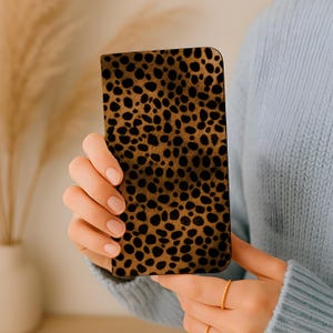 Peut inclure: Un étui de téléphone rectangulaire avec un motif léopard marron et noir. L'étui est tenu devant un pull bleu clair.