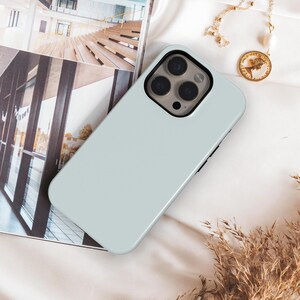 May include: A light sage green smartphone case with a triple camera cutout, featuring a smooth matte finish. The case is positioned on a surface next to a magazine showing an architectural interior and a gold chain necklace with a round pendant.
