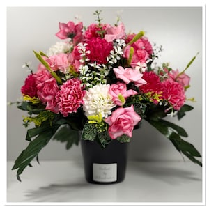 May include: A colourful floral display in a black vase. The arrangement includes pink, fuchsia, and white flowers, such as roses and carnations, with green leaves. The vase has a small white label with text.