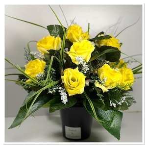 May include: A display of bright yellow roses in a black pot. The artificial flowers are set amongst green foliage and small white details. The pot has a label that reads "Handmade by Dawn Couture".