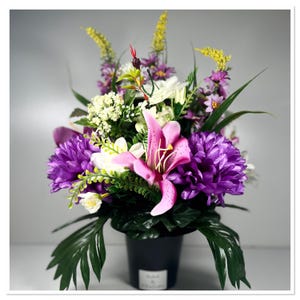 May include: A colourful floral display in a black pot. The arrangement includes a large pink lily, purple chrysanthemums, white flowers, and yellow accents. Green leaves provide a lush backdrop. The pot has a small white label.