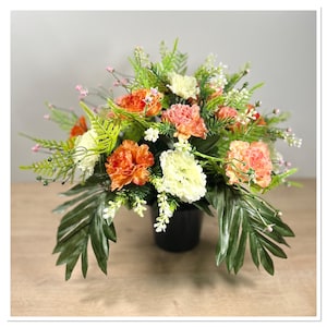 May include: A floral arrangement in a black pot with orange, white, and pink carnations and green foliage.