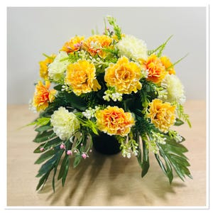 May include: A yellow and white artificial flower arrangement in a black pot. The arrangement features carnations and greenery.