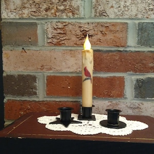 May include: Three black metal candle holders with a single white candle with a red cardinal design in the center holder. The candle is lit and the flame is visible. The candle holders are on a white lace doily on a dark brown surface.