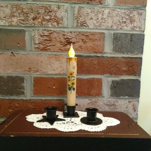 May include: A set of three candle holders with a lit candle in the center. The candle is decorated with sunflowers and has a black base. The candle holders are black metal and have a star-shaped base.