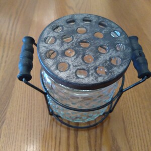 May include: Vintage glass flower frog with a metal lid and handles. The clear glass jar has a textured pattern. The lid has multiple holes for arranging flowers. The handles are black and the metal has a distressed finish.