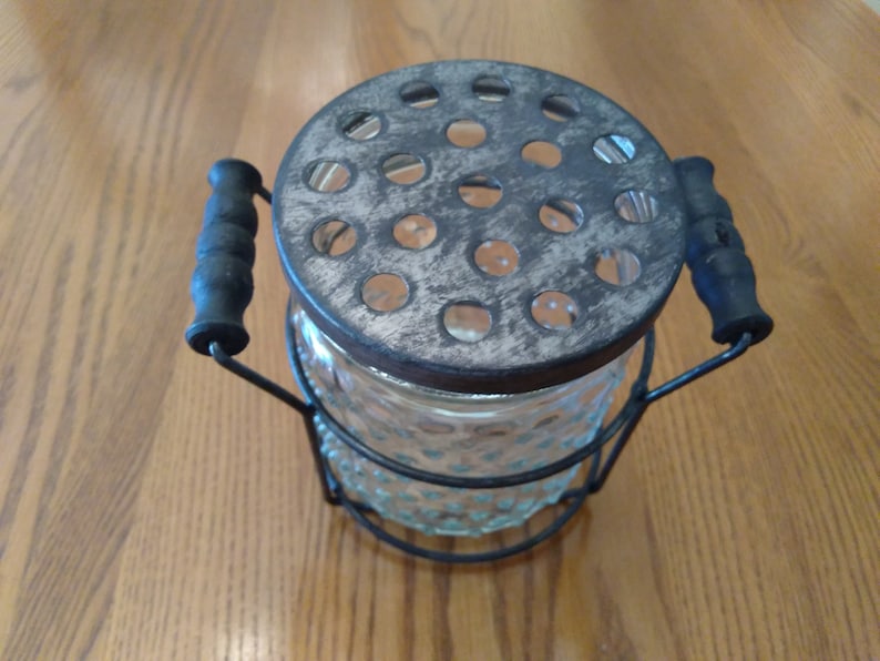 May include: Vintage glass flower frog with a metal lid and handles. The clear glass jar has a textured pattern. The lid has multiple holes for arranging flowers. The handles are black and the metal has a distressed finish.