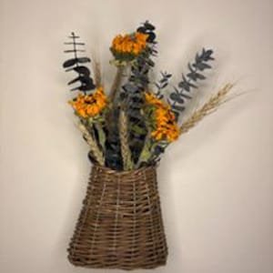 May include: A wall-mounted brown woven wicker basket containing a dried floral arrangement. The bouquet features bright orange sunflowers, stalks of golden wheat, and dark green eucalyptus leaves, creating a textured and natural display.