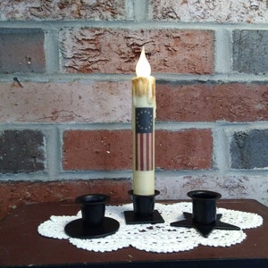 May include: Three black metal candle holders with a white candle with an American flag design in the center holder. The candle is lit and the holders are on a white doily.
