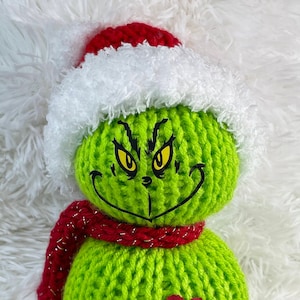 May include: A green knitted Grinch snowman wearing a red and white Santa hat with a red and silver scarf and a red heart on its chest.
