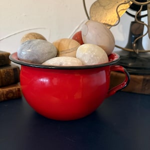 Puede incluir: Un cuenco de esmalte rojo brillante con borde y asa negros, lleno de piedras lisas y pulidas de varios colores. El cuenco se encuentra sobre una superficie azul oscuro, con un ventilador vintage al fondo.
