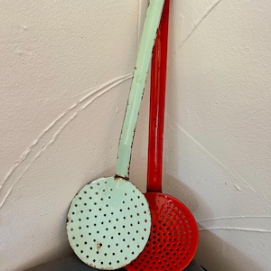 Pode incluir: Duas escumadeiras de cozinha vintage. Uma é verde menta, a outra é vermelha. Ambas têm tigelas redondas perfuradas e cabos longos. As escumadeiras estão encostadas a uma parede branca. A escumadeira verde menta está apoiada em um livro escuro.