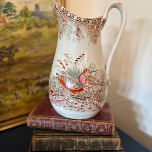 Può includere: Brocca vintage in ceramica bianca con un design dettagliato di uccelli e fogliame nei toni del rosso, arancione e verde. La brocca ha un bordo decorativo e un manico curvo. Poggia su una pila di libri antichi con copertine in pelle.