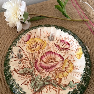 Op de afbeelding: Een rond decoratief bord met een centrale illustratie van gele en roze klaprozen met groen blad. Het bord heeft een geschulpte rand die is beschilderd in donkergroen met witte spikkels en een dunne gouden rand langs de binnenkant.