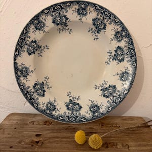 May include: A round ceramic plate featuring a white centre and a wide rim decorated with a dark blue floral pattern. The intricate design includes repeating clusters of flowers and scrolling foliage along the border, creating a detailed contrast.