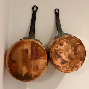 May include: Two copper saucepans with black handles are displayed against a white wall. The pans have a shiny, reflective surface and a rounded shape. The handles are long and have a loop at the end for hanging. The copper has a warm, reddish-brown hue.