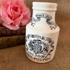 May include: A white ceramic Grey-Poupon mustard jar with a narrow neck and a wide base. The jar features black printed text and graphics, including the words Paris 1900, Maison Fondée en 1777, and Dijon. The design includes a central crest emblem.