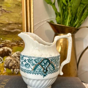 May include: A small, white ceramic creamer with a decorative teal and gold pattern. The creamer has a handle and a spout, and is trimmed with gold accents. The creamer is sitting on a dark surface.