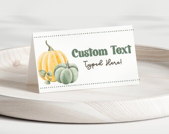 Op de afbeelding: Een wit gevouwen naamkaartje met een aquarelillustratie van een gele pompoen en een saliegroene pompoen. De kaart toont de tekst Custom Text Typed Here in groene en bruine letters, omlijst door twee horizontale stippellijnen.