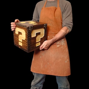 Puede incluir: Una caja de madera artesanal con acabado marrón oscuro y un gran signo de interrogación claro en la parte delantera. La caja tiene una tapa con bisagras y herrajes metálicos. Una persona con un delantal marrón sostiene la caja.