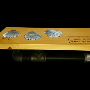Puede incluir: Una estantería de madera con tres conchas plateadas. La estantería tiene un soporte de tubería negro. El texto en la estantería dice: "Practice Good Hygiene Please Use The Three Seashells."