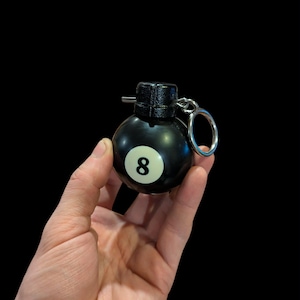 May include: A black novelty keychain in the shape of an eight ball grenade. The keychain has a white number eight on the black sphere, a black top, and a silver key ring. The background is black.