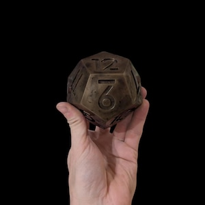 以下が含まれることがあります： ブロンズ色の12面ダイスで、1から12までの数字が面に刻まれています。黒い背景に、手でダイスが持たれています。6と12の数字が見えます。これは、テーブルゲームに使用される可能性があります。