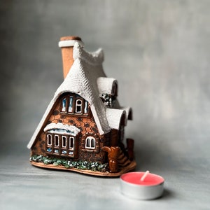 Può includere: Una casa in ceramica a forma di cottage con tetto bianco e muri in mattoni marroni. La casa ha un camino e finestre. C'è una piccola candela rossa davanti alla casa.