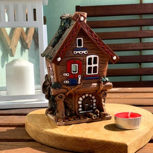 Peut inclure: Une figurine de maison en céramique avec un toit rouge, des bordures blanches et une porte rouge. La maison est décorée de vignes vertes et d'une cheminée. La figurine est posée sur une surface en bois.