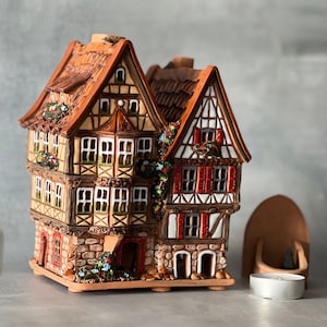 May include: A ceramic house shaped like a traditional German half-timbered building with red and white accents. The house has a brown roof and is decorated with green vines and flowers. The house is sitting on a base with a small candle holder.