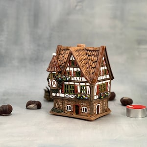 May include: A ceramic house figurine with a brown roof, white walls, and green trim. The house has a small tower on the side and windows with red shutters. The house is sitting on a brown base.